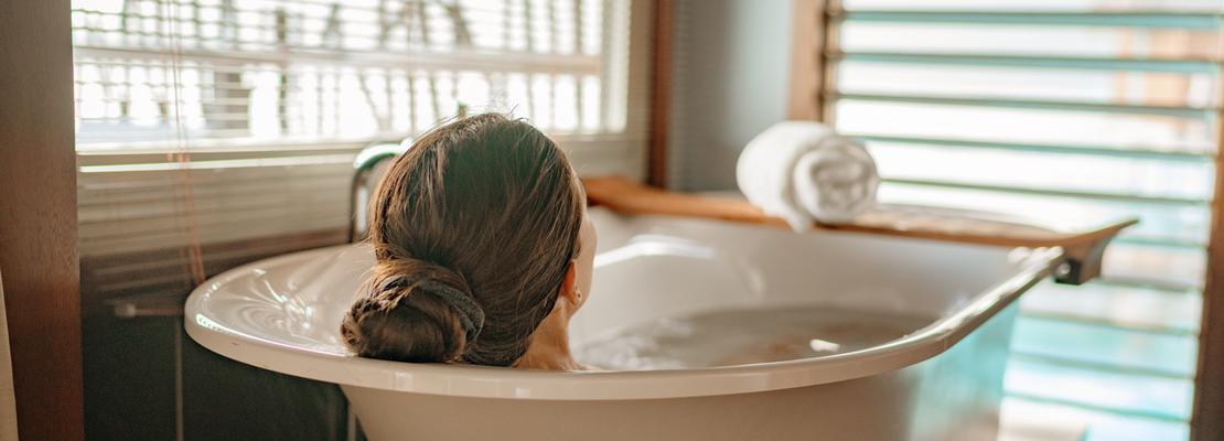 Rückansicht einer langhaarigen Person mit dunkelblondem Haar, die entspannt in einer freistehenden Badewanne liegt.